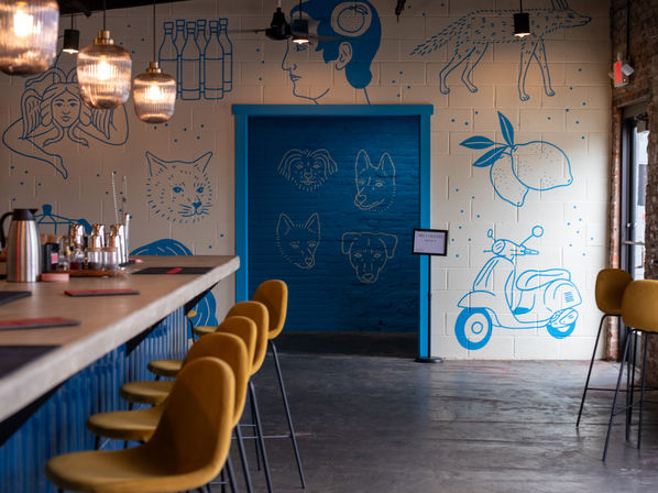 Modern industrial-style bar interior with mustard-yellow stools, hanging pendant lights and a playful blue mural on white cinderblock walls showing animals, lemons and a scooter