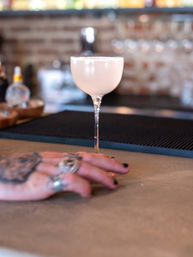 Pale pink frothy cocktail in a coupe glass on a bar counter, with a blurred tattooed hand wearing rings in the foreground and a warm brick bar background.