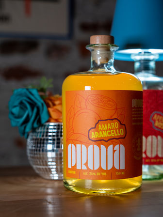 Bright orange citrus amaro liqueur bottle with wooden stopper on a wooden table, teal flower and blue lamp blurred in the background