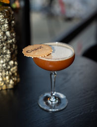 Espresso-brown cocktail in a coupe glass with a creamy foam top and a round biscuit stamped "Ciao!" perched on the rim, served on a dark wood bar table