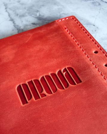 Close-up of a vibrant red leather journal cover with bold embossed lettering, neat stitched spine and punched holes, photographed on a light marble tabletop.
