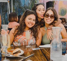 Three friends enjoying a sunny outdoor brunch patio, smiling and posing with mimosas and carafes on a wooden table with a burger, utensils, tropical plants and a colorful mural in the background.