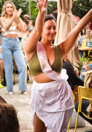 Bride-to-be dancing with arms raised at an outdoor bachelorette brunch on a lively patio, wearing a pink "Bride" sash, olive crop top and white wrap skirt while friends applaud.