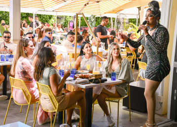 Sunny outdoor brunch patio with a drag performer entertaining a lively crowd seated on yellow chairs around tables filled with mimosas, pitchers and brunch plates.