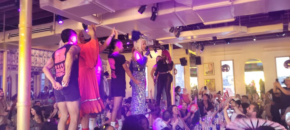 Vibrant drag brunch in a downtown bar with a sequined performer and dancers standing on tables under purple lights as a cheering crowd claps and takes photos