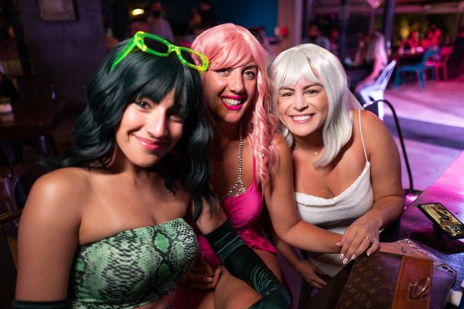 Three smiling friends in colorful wigs and party outfits pose at a neon-lit nightclub bar, laughing together over a table with a purse and phone — lively city nightlife scene.
