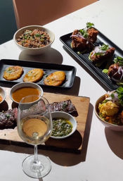 Sunlit restaurant table with grilled steak on a wooden board served with chimichurri and dipping sauces, crispy fried chicken bites with red onion and cilantro on a black plate, three golden tostones, bowls of rice and vegetable salad, and a glass of white wine.
