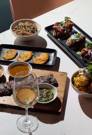 Sunlit restaurant table with grilled steak on a wooden board served with chimichurri and dipping sauces, crispy fried chicken bites with red onion and cilantro on a black plate, three golden tostones, bowls of rice and vegetable salad, and a glass of white wine.