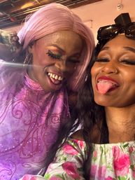 Close-up party selfie of a drag performer in a pink wig and sparkly purple dress with colorful eye makeup laughing beside a friend in a floral off-shoulder top sticking out her tongue and wearing sunglasses on her head.