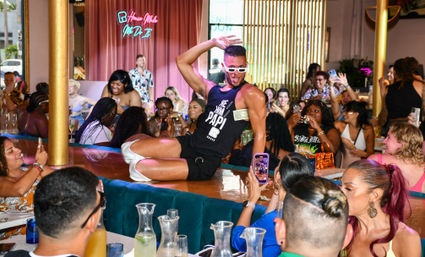 Energetic male performer in sunglasses and a 'Papi' tank posing on a bar while a lively brunch crowd snaps photos and cheers in a colorful indoor restaurant with neon accents.