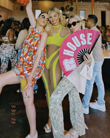 Three women posing at a lively indoor daytime party: left in a bright soda-can print mini dress holding a mimosa, center in a neon lime cutout bodysuit, right wearing sunglasses and patterned flared pants while holding a large pink fan that reads HOUSE.