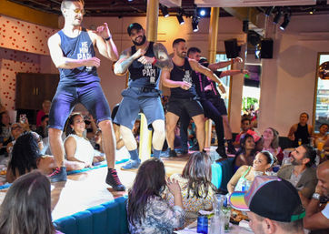 High-energy male dancers in matching tank tops performing on a raised stage inside a lively restaurant, entertaining a seated, smiling crowd who cheer, clap and take photos.