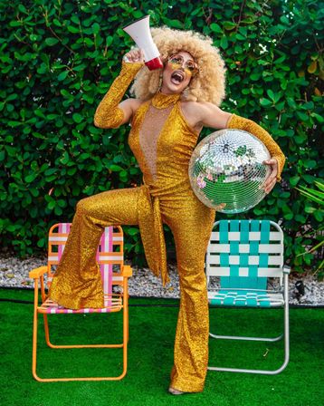 Energetic performer in a gold sequin 1970s-style jumpsuit and big curly wig, holding a megaphone and a mirrored disco ball while posing between colorful lawn chairs in a lush backyard garden party scene.