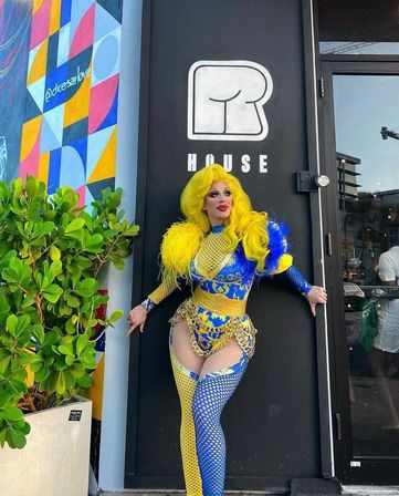 Drag performer in a bright yellow wig and blue-and-yellow ornate costume poses at an urban storefront doorway beside a colorful geometric mural and potted greenery.