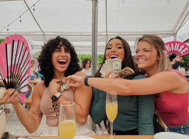 Three laughing friends at an outdoor brunch under a white canopy — one with a pink hand fan, another holding a stack of cash while a friend tucks a dollar into her top, with mimosas on the table.