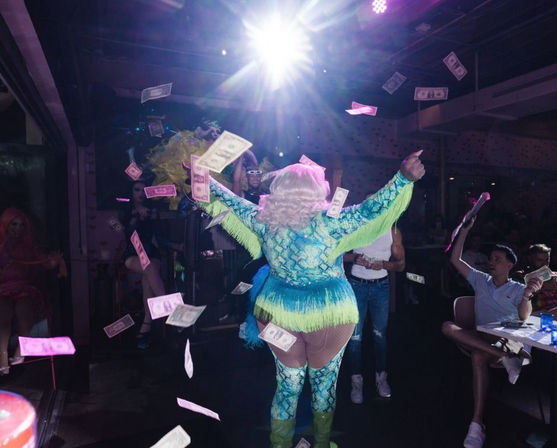 Drag performer in turquoise fringe costume and blonde wig on a nightclub stage with arms raised as dollar bills rain down and an audience at tables cheering.