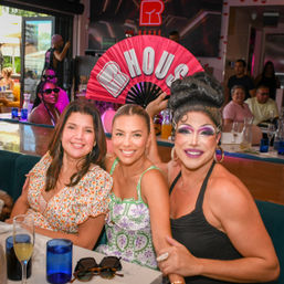 Smiling trio at a lively daytime brunch: two friends and a glamorous drag performer in bold makeup holding a pink fan that reads "HOUSE".