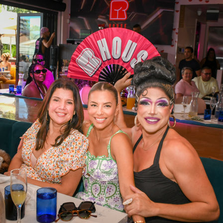 Smiling trio at a lively daytime brunch: two friends and a glamorous drag performer in bold makeup holding a pink fan that reads "HOUSE".