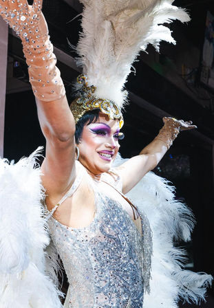 Cabaret performer in showgirl costume: glittering silver sequin dress, tall white feather headdress and wings, bold purple eye makeup and rhinestone gloves, arms raised and smiling on stage.