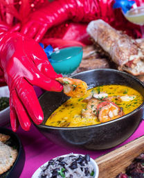 Glossy red-gloved hand picking a shrimp from a creamy yellow seafood stew in a black bowl, surrounded by black beans and rice, crusty bread and a blue cocktail in the background