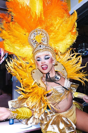 Smiling drag performer in a towering yellow feathered headdress and gold sequined costume, dramatic makeup and energetic pose on a nightclub/cabaret stage