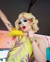 Drag performer on stage in a wavy platinum-blonde wig, dramatic blue eye makeup and red lipstick, wearing a bright yellow sequined feathered costume, sparkly bracelet and holding a microphone with one arm raised revealing a tattoo.
