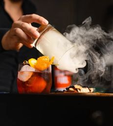 Smoky craft cocktail on a dark bar as smoke is poured from a glass over an ice-filled amber drink garnished with orange peel.