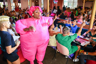 Drag performer in an inflatable hot-pink costume entertains a lively drag-brunch crowd in a colorful restaurant as guests cheer, film on phones and sip champagne.