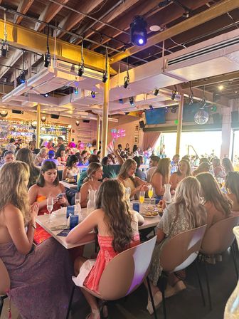 Trendy weekend brunch crowd at a lively downtown restaurant — friends at communal tables sipping mimosas, colorful lighting, exposed wooden beams, neon wall sign and busy bar in the background.