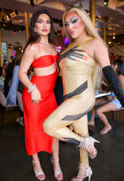 Two glamorous drag performers posing at a vibrant lounge: one in a red cutout dress and silver heels, the other in a gold rhinestone catsuit with dramatic makeup and strappy crystal heels.