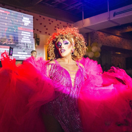 Drag performer in a glittering hot-pink gown with voluminous tulle sleeves, dramatic makeup and curly wig, striking a pose on a lively nightclub stage during a drag show.