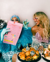 Two glamorous drag performers at a colorful drag brunch — one in blue sequins and feathers peeking over a pink menu and holding a champagne flute, the other in a pink sparkly top and heart-shaped sunglasses smiling over a platter of croissant sliders topped with disco-ball picks.