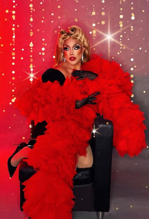 Glamorous retro performer in a voluminous red feather boa and black gloves, seated on a black chair wearing blonde vintage curls, dramatic makeup, fishnet stockings and black stiletto heels against a sparkling red bokeh studio backdrop.