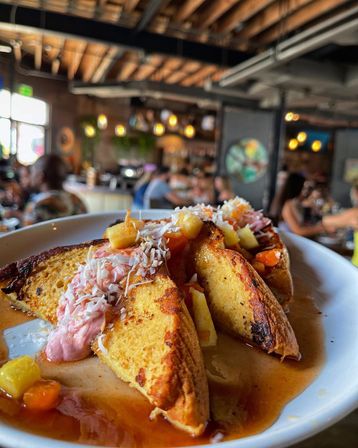 Thick golden French toast slices topped with pink cream, shredded coconut, diced pineapple and syrup on a white plate in a busy rustic cafe with exposed wooden beams.