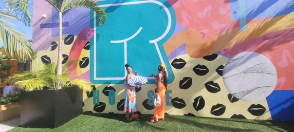 Two people posing on green turf in front of a colorful outdoor mural with a large teal letter, black lip prints, pink and orange shapes, a marble-like circle, and palm plants