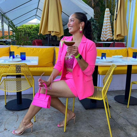 Person in bright pink blazer and matching shorts holding a cocktail and tiny pink purse, seated at a sunny outdoor patio cafe with yellow cushions, striped umbrellas and blue glassware.