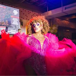 Drag performer in a sparkly hot-pink fringe dress and voluminous tulle cape, dramatic makeup and curly wig, striking a pose on an indoor nightclub stage during a lively drag show.