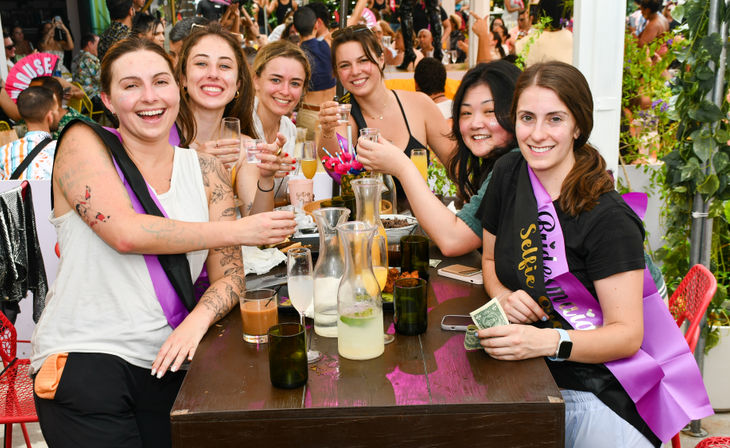 Group of women smiling and toasting at a lively outdoor patio bachelorette brunch, pitchers, cocktails and snacks on the table