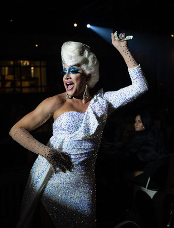 Drag performer in a sparkling white one-shoulder gown and white wig, dramatic blue eye makeup and jeweled gloves, holding dollar bills aloft under a spotlight during a lively nightclub cabaret performance.