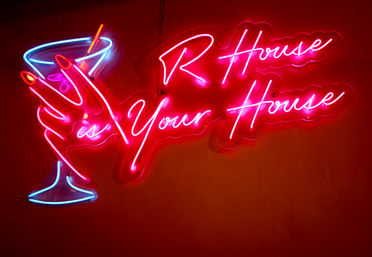 Vibrant pink-and-blue neon bar sign of a martini glass with cursive text reading is Your House, glowing on a red wall