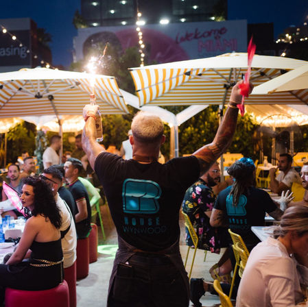 Miami outdoor patio at night: server raises a bottle with a lit sparkler while guests dine under striped umbrellas and string lights