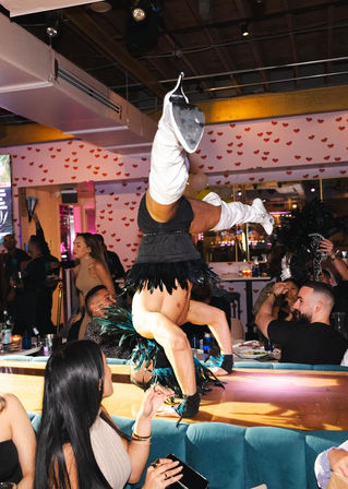 Cabaret performer doing a handstand on a dining table in a lively nightclub, wearing white knee-high boots and a feathered costume while diners watch under heart-patterned wallpaper.
