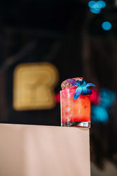 Bright red-orange tropical cocktail in a short glass with crushed ice, topped with a blue orchid and dried citrus, set on a pale ledge against blurred neon bar lights.