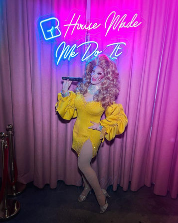 Drag performer in a bright yellow beaded mini dress with puffed sleeves, fishnet tights and sparkly heels holds a microphone and poses in front of pink curtains and a neon sign reading "House Made Me Do It".