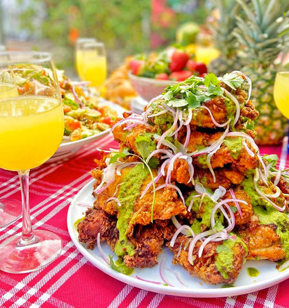 Stack of crispy fried chicken drizzled with bright green herb sauce, topped with pickled red onions and cilantro on a white plate at a sunny outdoor brunch table with mimosas, salad and pineapple on a red checkered cloth.