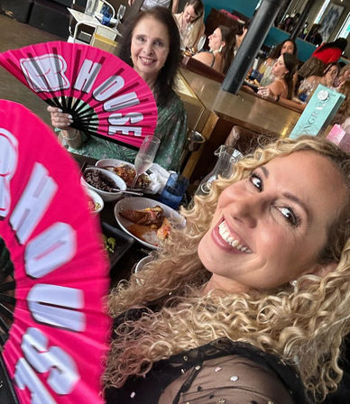 Two smiling women taking a selfie at a lively indoor restaurant brunch, holding bright pink hand fans with plates of food and champagne glasses on the table.