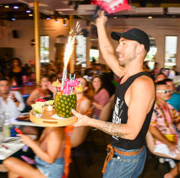 Server carrying a sparkler-topped pineapple cocktail on a wooden tray through a crowded, lively tiki-style brunch restaurant.