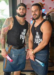 Two bearded men posing in a colorful indoor bar, wearing black "HE/HIM PAPI" tank tops and denim aprons, showing tattoos and holding folded umbrellas — casual bartenders/servers.