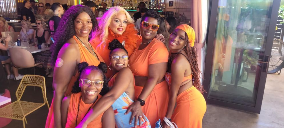 Six smiling women in coordinated orange outfits with a glamorous performer in a feather boa, posing inside a lively sunlit bar/restaurant with tables and patrons in the background.
