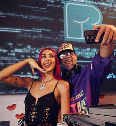 Playful selfie of a woman with pink hair and a DJ wearing headphones at a colorful nightclub DJ booth, turntables and vibrant lights in the background.